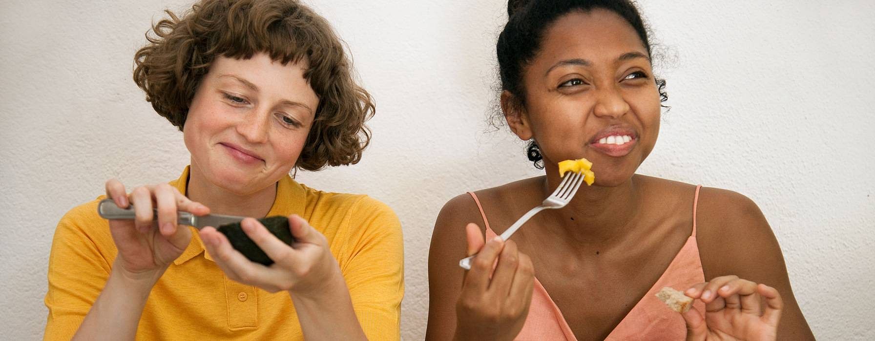 due ragazze: una mangia un avocado con un coltello, l'altra l'ananas con una forchetta