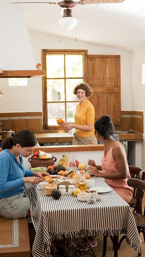 tre ragazze in cucina, due sedute al tavolo che mangiano