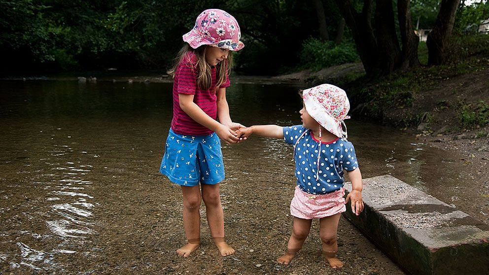 Bambino e bambina più grande con cappellini e piedi nel laghetto si scambiano qualcosa tra le mani