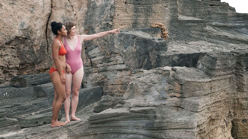 Due ragazze in costume su una roccia indicano qualcosa in lontananza